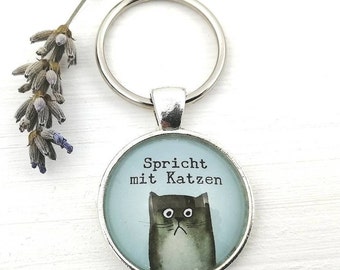 May include: A silver-colored keychain with a round pendant featuring a watercolor illustration of a gray cat with the text "Spricht mit Katzen" on a light blue background. A sprig of lavender is in the background.