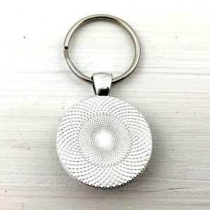 Puede incluir: Un llavero plateado con un colgante redondo. El colgante presenta un dise&ntilde;o en espiral blanco y gris. El anillo del llavero est&aacute; unido al colgante con un peque&ntilde;o lazo de metal.