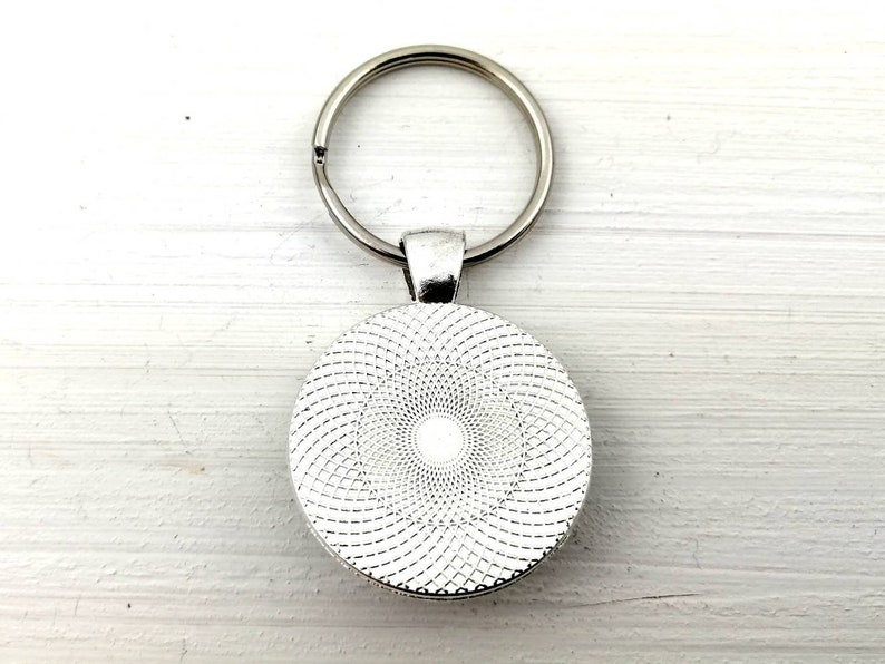 Puede incluir: Un llavero plateado con un colgante redondo. El colgante presenta un dise&ntilde;o en espiral blanco y gris. El anillo del llavero est&aacute; unido al colgante con un peque&ntilde;o lazo de metal.