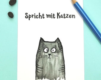 May include: A white card with the German text "Spricht mit Katzen" and a cartoon cat illustration in shades of gray. A blue pencil and two coffee beans are also visible.
