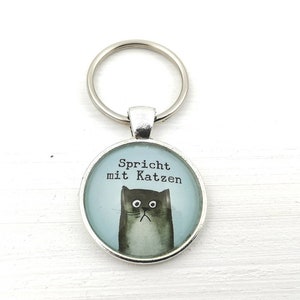 Puede incluir: Llavero plateado con un colgante redondo. El colgante muestra una ilustraci&oacute;n en acuarela de un gato gris y el texto "Spricht mit Katzen" sobre un fondo azul claro. El gato tiene una expresi&oacute;n triste.