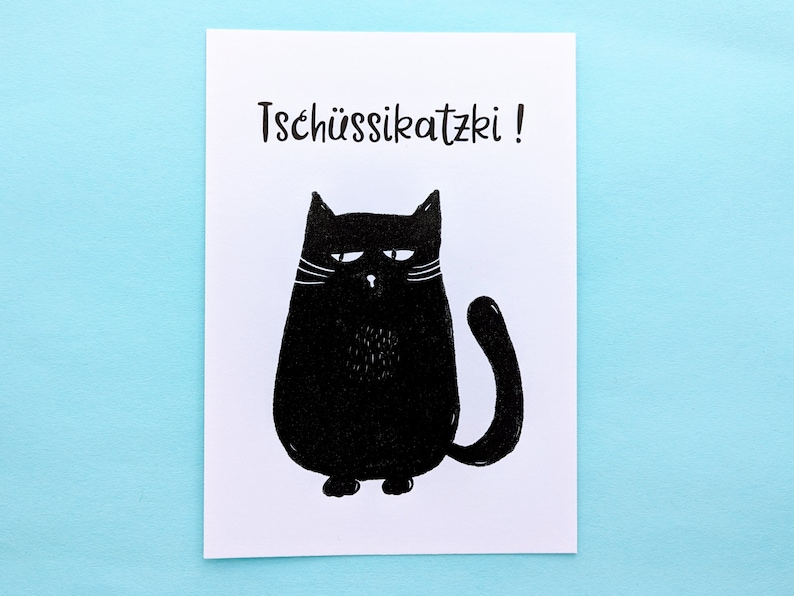 May include: A white card with a black illustration of a grumpy cat. The cat has a round body, a curved tail, and white whiskers. The text "Tsch&uuml;ssikatzki!" is printed above the cat in black.