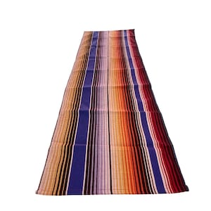 May include: A long, narrow, striped table runner with alternating bands of orange, brown, purple, and blue. The runner is made of a woven fabric and has a slightly textured surface.