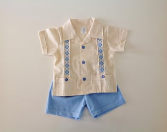 May include: A two-piece outfit for a baby or toddler. The shirt is cream colored with blue and white embroidered trim. The shorts are light blue.