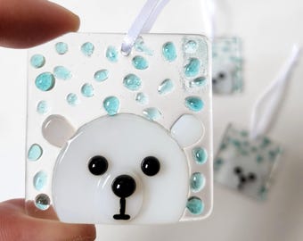 May include: Square, clear glass ornament featuring a white polar bear face with black eyes and nose. The ornament has blue glass accents and a white ribbon for hanging. Additional ornaments are visible in the background.