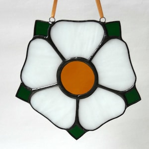 May include: White stained glass flower with a yellow center and green accents. The flower is framed in black and has a white background.