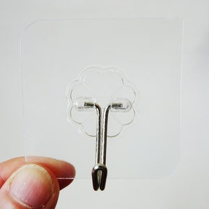 May include: A clear plastic adhesive hook with a silver metal hook. The hook is designed to be attached to a smooth surface.