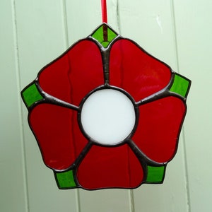 May include: A red stained glass flower with a white center and green accents. The flower is hanging from a red ribbon.