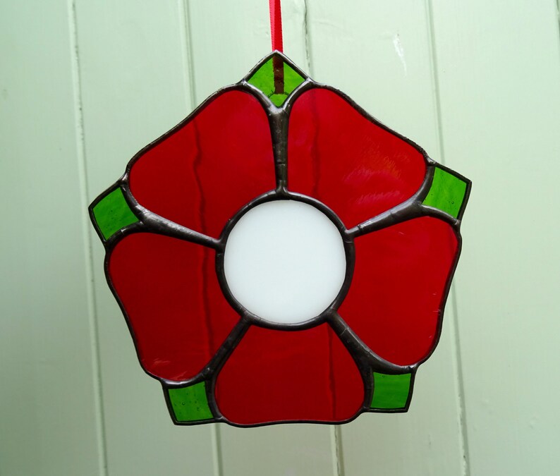 May include: A red stained glass flower with a white center and green accents. The flower is hanging from a red ribbon.
