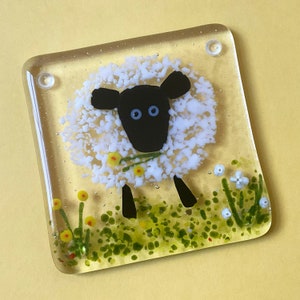 May include: A glass coaster featuring a white sheep with black face and legs, surrounded by green and yellow flowers. The coaster is made of clear glass with a textured surface.