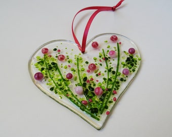 May include: A heart-shaped glass ornament with a floral design. The ornament features green and pink accents on a white background. A red ribbon is attached for hanging. The ornament is approximately 4 inches wide.