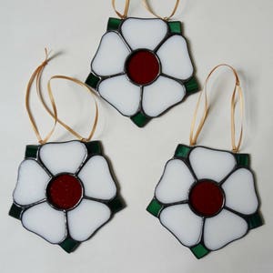 May include: Three white stained glass ornaments shaped like flowers with red centers and green outlines. Each ornament has a gold ribbon attached for hanging.