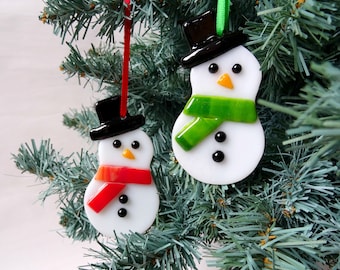 Op de afbeelding: Twee sneeuwpopornamenten hangen aan een kerstboom. Elke sneeuwpop is wit met een zwarte hoge hoed, zwarte knoopogen en een wortelneus. De ene heeft een rode sjaal, de andere een groene sjaal. Feestelijke kerstversieringen.