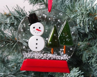 Puede incluir: Adorno navideño de cristal con forma de globo de nieve. Presenta un muñeco de nieve con sombrero de copa negro, dos árboles verdes y una base roja. El adorno es transparente con detalles blancos y una cinta roja para colgar.