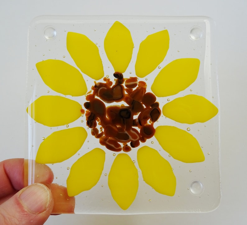 Peut inclure: Un dessous de verre en verre avec un motif de tournesol. Le tournesol est jaune avec un centre brun. Le dessous de verre est transparent et de forme carr&eacute;e.