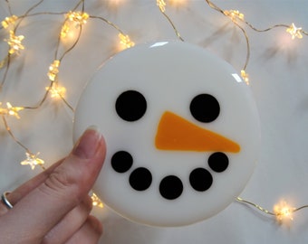 Op de afbeelding: Een rond, wit sneeuwpopgezicht met zwarte ogen en mond, en een oranje wortelneus. De sneeuwpop wordt in een hand gehouden, met een slinger van stervormige lampjes op de achtergrond.