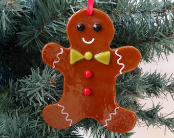Op de afbeelding: Een glanzende bruine peperkoekman kerstversiering. Het heeft zwarte ogen, een witte glimlach, een groene strik, rode knopen en witte glazuurdetails. De versiering hangt aan een rood lint.