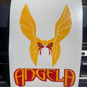 May include: A white rectangular decal featuring a stylized yellow winged helmet graphic with red and green accents. Below the helmet, the name ANGELA is printed in bold red capital letters with a yellow outline on a white background.