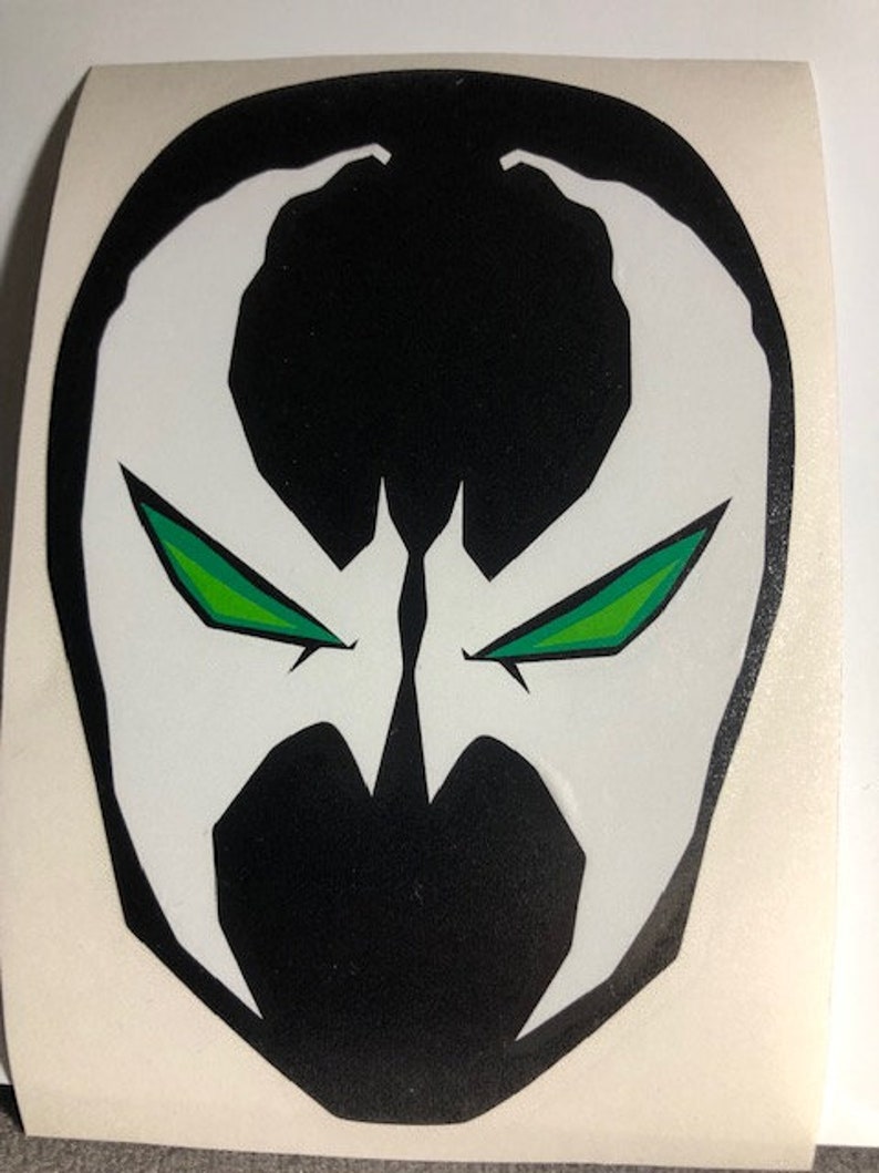 May include: A sticker featuring the Spawn character's mask. The mask is primarily black with white accents and sharp, angular shapes. The eyes are green with a gradient effect, adding a striking contrast to the design.