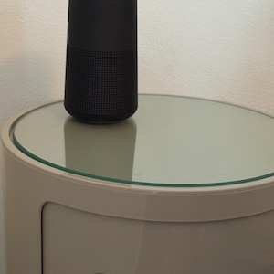 May include: A black cylindrical speaker with a perforated lower section and a solid top. The speaker is placed on a round, glass-topped table. The table is supported by a white, rounded cabinet with a black circular opening.
