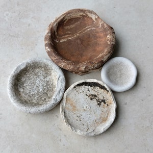 May include: An overhead view of four small, irregularly shaped stone bowls. The largest bowl is a deep brown colour, while the others are shades of grey and white. The bowls have rough, textured surfaces, suggesting a natural, handcrafted aesthetic.