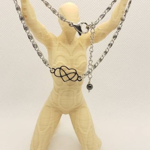 May include: A silver chain necklace with a black heart-shaped pendant featuring a knot design. The necklace is displayed on a mannequin.