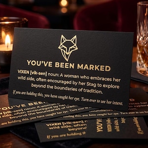 May include: Black cards with gold text that read "YOU'VE BEEN MARKED" and a fox logo. The cards are on a dark wooden table with a candle and a glass of whiskey in the background. The text defines "VIXEN".