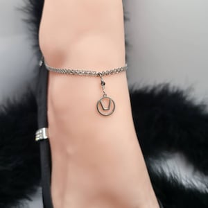 Puede incluir: Un brazalete de tobillo plateado con un pequeño colgante circular con un diseño geométrico. El brazalete está en el tobillo de una persona.