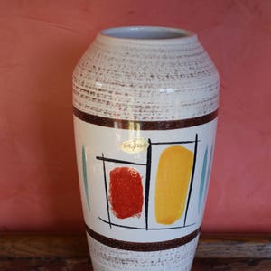 Könnte beinhalten: Weiße Keramikvase mit geometrischem Design mit roten, gelben und blauen Akzenten. Die Vase hat einen braunen Streifen oben und unten. Die Vase ist mit "Schirnding" in goldenen Buchstaben beschriftet.