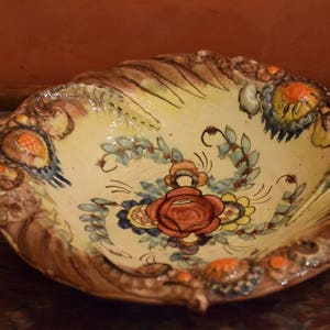 Könnte beinhalten: Eine dekorative Keramikschale mit einem gewellten Rand und einem floralen Design in Orange-, Gelb-, Blau- und Grüntönen. Die Schale hat ein rustikales, handbemaltes Aussehen.