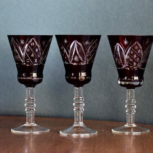Op de afbeelding: Drie robijnrode wijnglazen met ingewikkelde geëtste ontwerpen. De glazen hebben een heldere, gedetailleerde steel en voet. De geëtste patronen omvatten diamant- en stervormen. De glazen worden gepresenteerd op een houten oppervlak tegen een blauwgroene achtergrond.