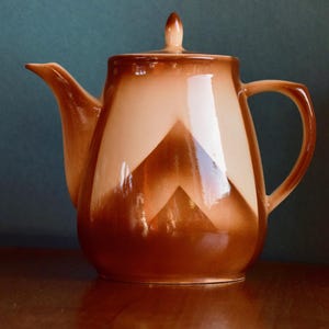 Könnte beinhalten: Eine braun-beige Keramik-Teekanne. Die Teekanne hat einen Henkel, einen Ausguss und einen Deckel mit einem kleinen Knopf. Das Design zeigt ein geometrisches Muster auf der Vorderseite. Die Teekanne steht auf einer Holzoberfläche.