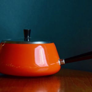 Op de afbeelding: Oranje fonduepan met een zwarte handgreep en een zilveren deksel. De pan staat op een houten oppervlak.
