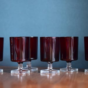 Könnte beinhalten: Sechs klare, gestielte Trinkgläser mit einer dunkelroten Tönung. Die Gläser sind in einer Reihe auf einer Holzoberfläche angeordnet.