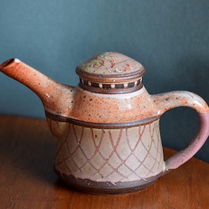 Könnte beinhalten: Eine Keramik-Teekanne mit Deckel, Ausguss und Henkel. Die Teekanne hat eine gesprenkelte, strukturierte Oberfläche mit Farbtönen von Beige, Orange und Braun. Der Körper weist ein Kreuzschraffurmuster in Rot auf. Der Griff ist rosa.