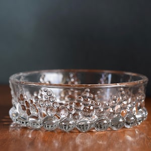 Könnte beinhalten: Eine transparente Glasschale mit einem strukturierten, blasenartigen Muster an den Seiten. Die Schale ist kreisförmig und steht auf einer Holzoberfläche, mit einem dunklen Hintergrund. Das Design verleiht der Schale ein einzigartiges, dekoratives Aussehen.