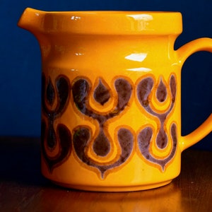 Könnte beinhalten: Eine gelbe Keramikkanne mit einem braunen und orangefarbenen geometrischen Muster. Die Kanne hat einen Henkel und einen Ausguss.