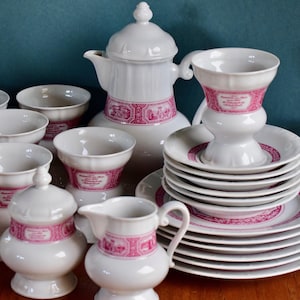 Op de afbeelding: Een wit porseleinen theeservies met roze decoratieve banden. De set bevat een theepot, kopjes, schotels, een melkkan en een suikerpot. De kopjes en schotels hebben een geschulpte rand.
