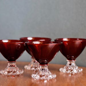 Könnte beinhalten: Vier Vintage-Cocktailgläser in Rot. Jedes Glas hat eine konische Schale, einen klaren Stiel mit rotem Band und eine Basis aus klaren Glasblasen. Die Gläser sind auf einer Holzoberfläche angeordnet.