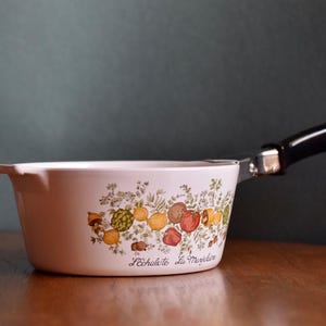 May include: White ceramic saucepan with a black handle and a decorative vegetable and herb design. The pan features a curved shape and a small spout. The text "L'échalote La Marjolaine" is printed on the side.