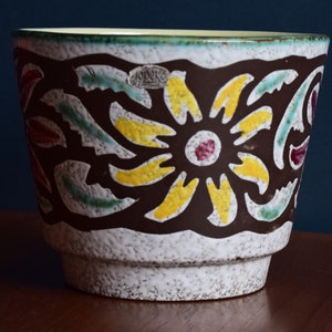 Könnte beinhalten: Ein weißer Keramiktopf mit einem braunen, gelben, roten und grünen Blumendesign. Der Topf hat eine strukturierte Oberfläche und ein Etikett mit der Aufschrift "JUNK" in einem blauen Oval.