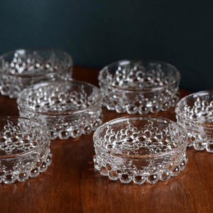 Könnte beinhalten: Sechs klare Glasschalen mit einem strukturierten, blasenartigen Design. Die Schalen sind in einem Halbkreis auf einer Holzoberfläche angeordnet.