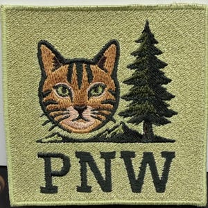 May include: A square embroidered patch with a light olive green background. The design features a brown tabby cat head, a dark green pine tree, and a small mountain silhouette. The letters PNW are stitched in large, bold, dark green font below.