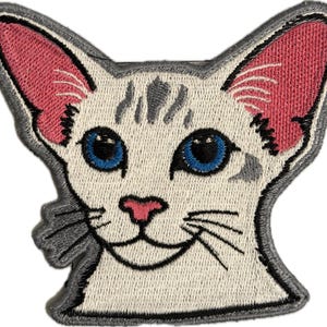 May include: Embroidered patch featuring a cat's face with blue eyes, pink ears, and a pink nose. The cat has gray markings on its head and whiskers. The patch is outlined in gray.