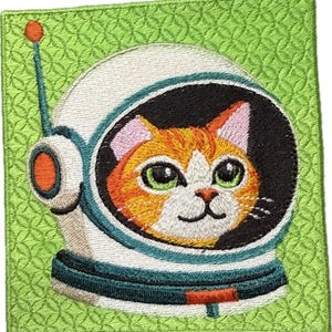 May include: Embroidered patch featuring a cat wearing an astronaut helmet. The cat has orange and white fur with green eyes and pink ears. The helmet is white with teal and black accents. The patch is on a green background.