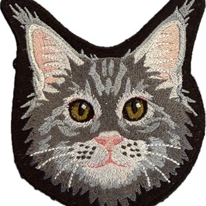 May include: An embroidered patch featuring the head of a grey tabby cat with prominent ear tufts. The cat has golden-brown eyes, a light pink nose, and white whiskers. The design is set against a solid black background with a shaped border.