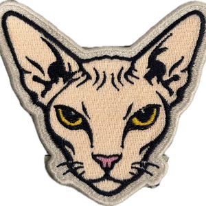 May include: Embroidered patch featuring a Sphynx cat's face. The cat has a light beige complexion, yellow eyes, a pink nose, and black details. The patch is outlined with a light gray border.