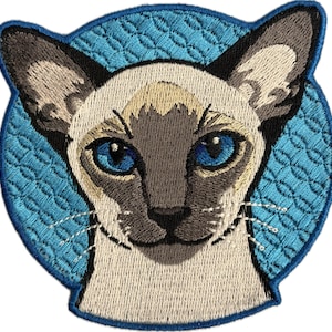 May include: Embroidered patch featuring a Siamese cat's face. The cat has blue eyes, tan fur, and dark brown ears and muzzle. The patch is set against a light blue, textured background with a scalloped edge.