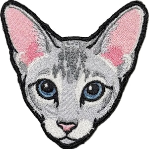 May include: An embroidered patch featuring the head of a cat with light gray fur, dark gray tabby markings on the forehead, large pointed ears with pink inner coloring, and bright blue eyes. The patch has a thick black stitched border.