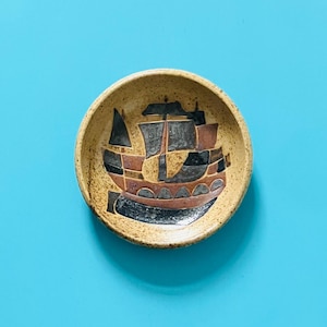 May include: A round ceramic bowl with a speckled beige finish featuring a hand-painted graphic of a sailing ship. The ship design incorporates geometric shapes in shades of black, brown, and grey, centred within the interior of the bowl.